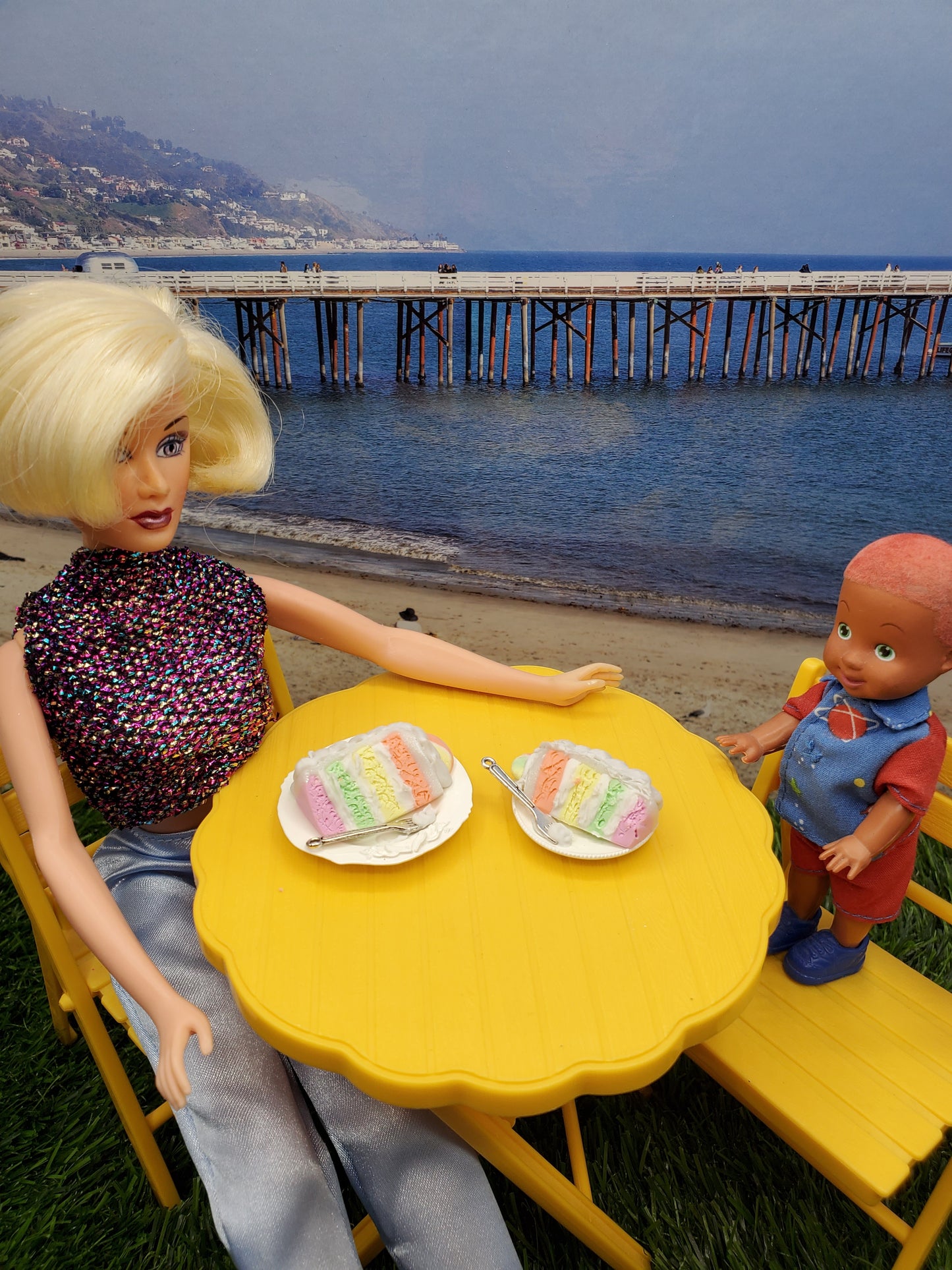 Fashion doll enjoying a sherbet cake with a child at a yellow table by the beach