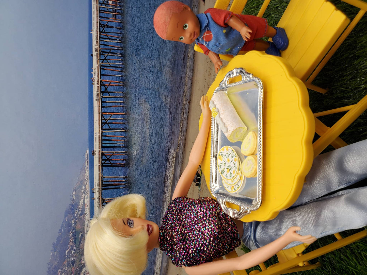 Barbie doll serving lemon sponge rolls for dolls on a yellow table with a child doll by the beach