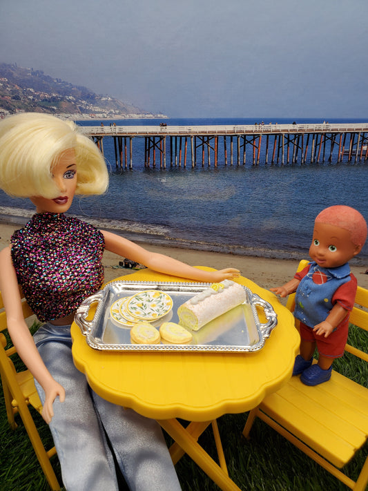 Barbie doll serving lemon sponge rolls for dolls on a yellow table with a child doll by the beach