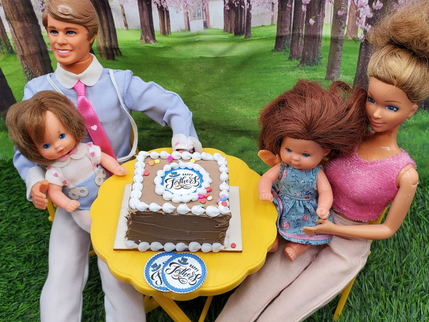Miniature Father's Day cake on a table with dolls celebrating outdoors in a park setting