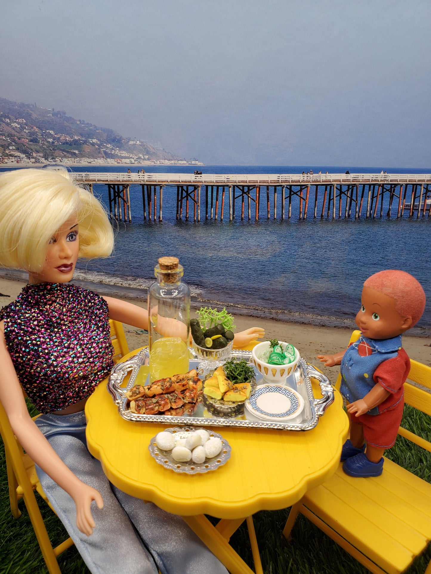Chef Gina's miniature Greek food set displayed on a table by the beach, featuring vibrant dishes and drinks.