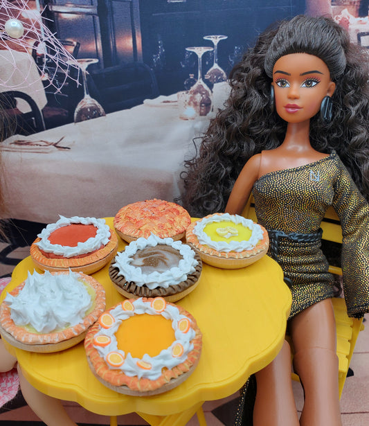 Miniature lemon meringue pie and assorted mini pies on yellow table next to doll in sparkly dress