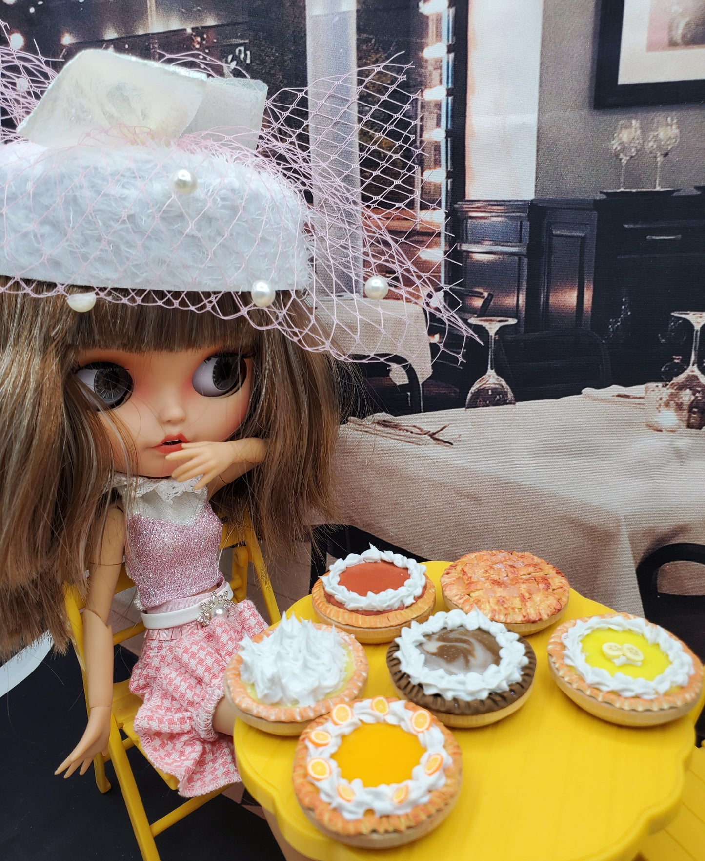 Miniature orange cream pie and other mini pies displayed on a yellow table next to a doll in a pink outfit with a white hat