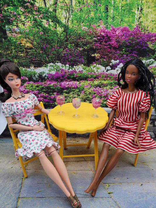 Two dolls sitting at yellow table with three miniature pink lime doll drink glasses in garden setting