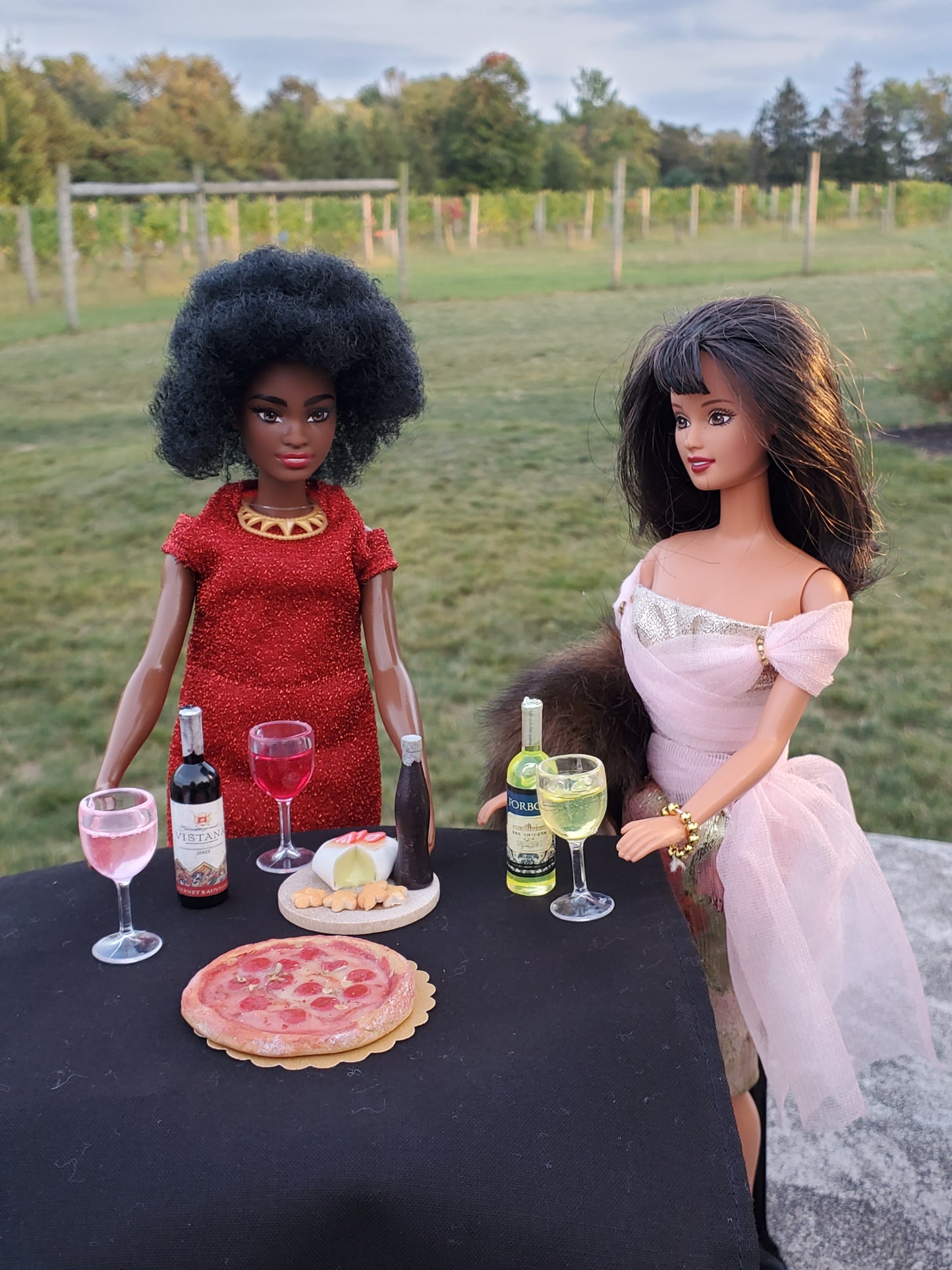 Miniature pink wine glass for dolls displayed with two dolls and food accessories on a black table outdoors