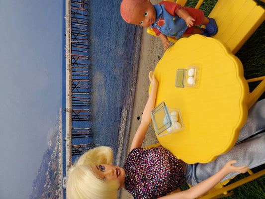 Barbie doll and child doll at a beach table displaying miniature rum balls for dolls