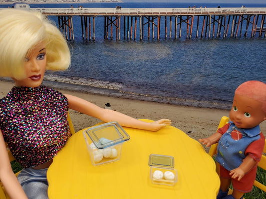 Barbie doll and child doll at a beach table displaying miniature rum balls for dolls