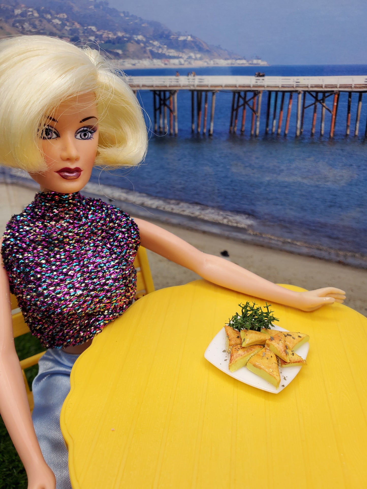 Barbie doll enjoying miniature spanakopita fashion dolls on a beachside table with a scenic view