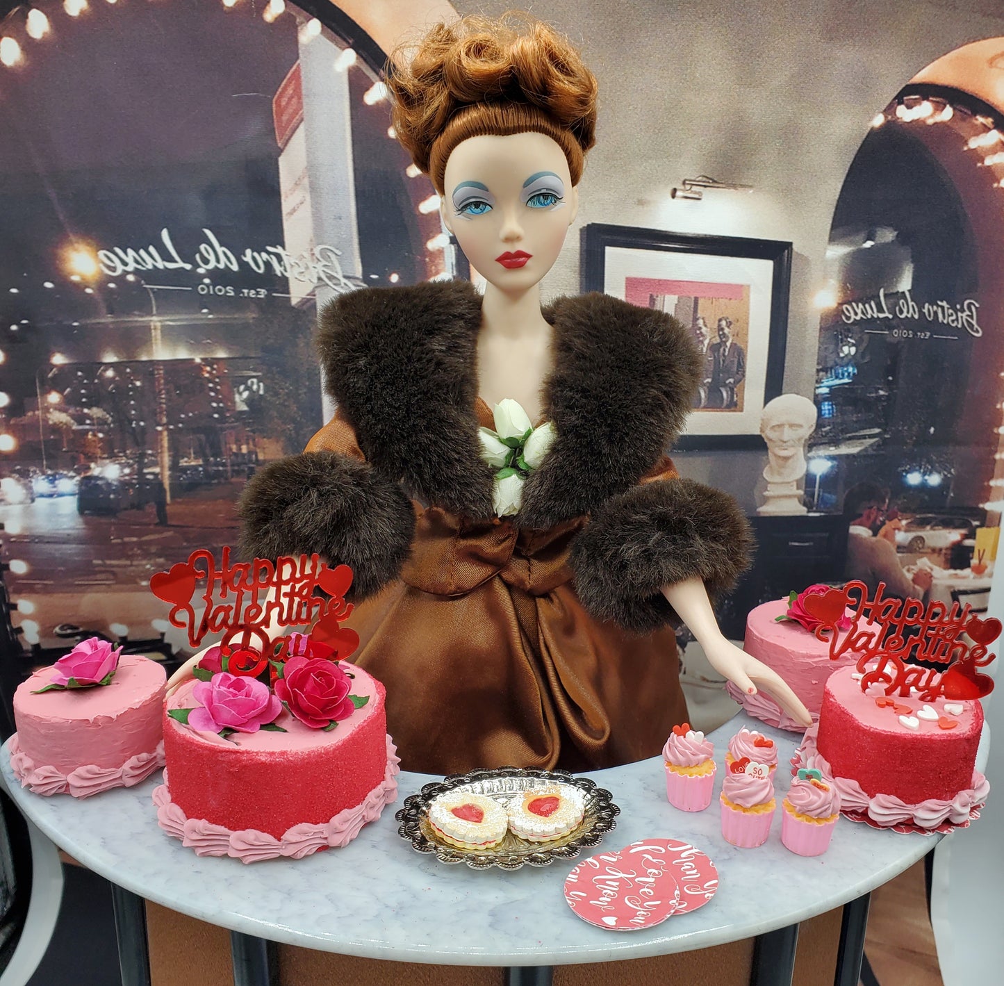 Miniature Valentine's cupcakes and cakes displayed with a doll dressed in brown fur on a marble table for Valentine's Day