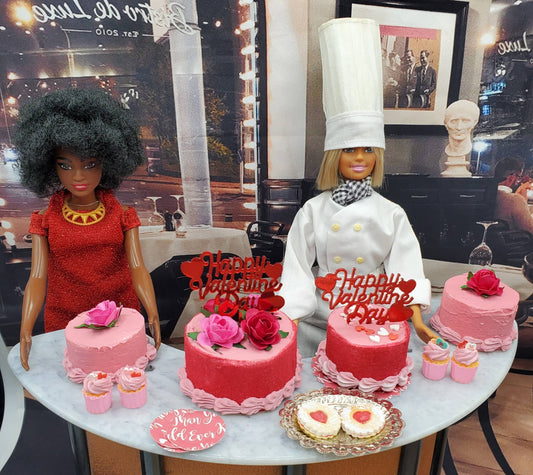 Miniature Valentine's Day cake and cupcakes displayed with dolls dressed as baker and customer on table