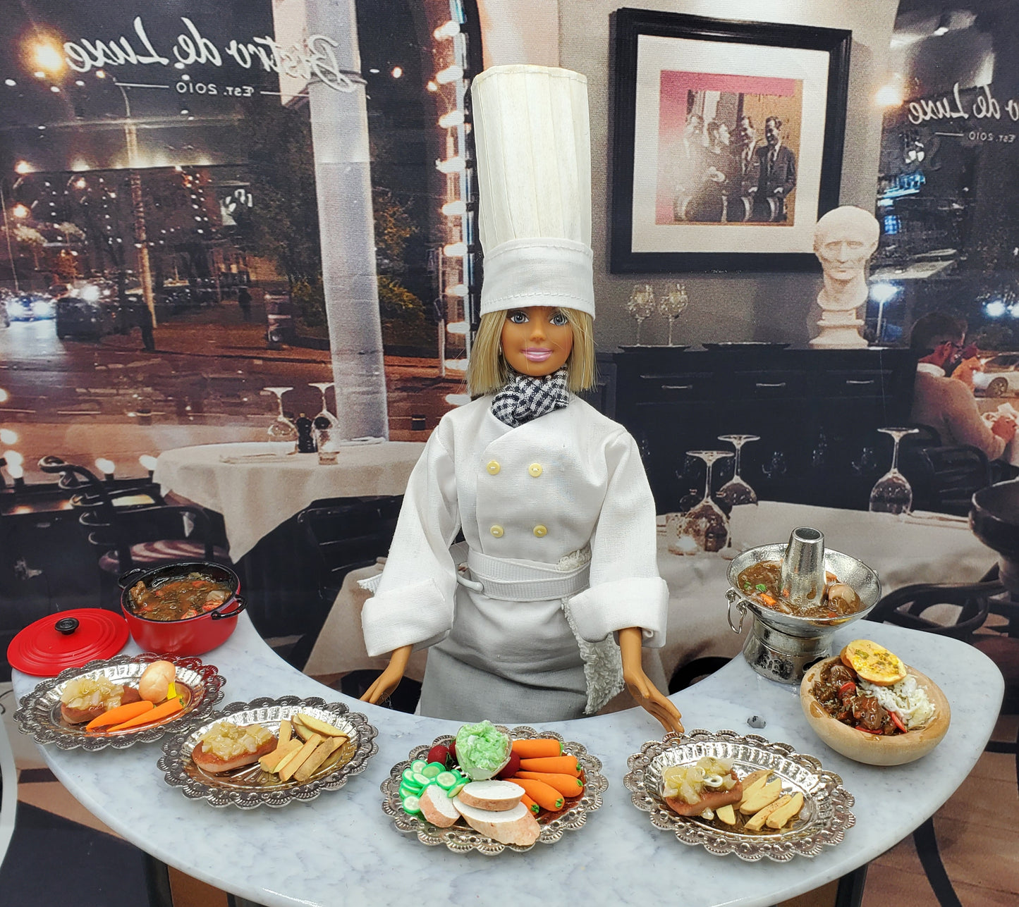 Chef doll dressed in white with a tall hat presenting a miniature veggie platter and assorted mini dishes on a marble counter