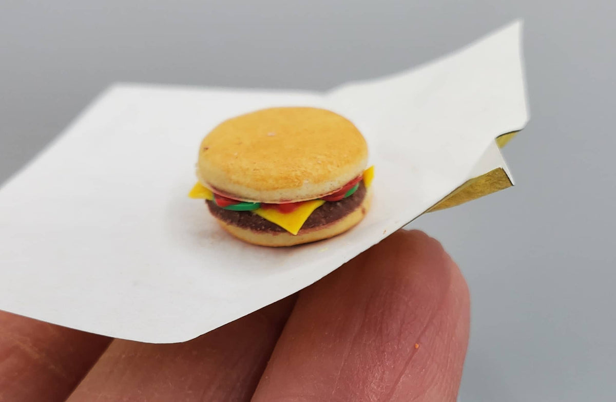 A miniature toy hamburger on a piece of white paper, held between a person's fingers against a neutral gray background.