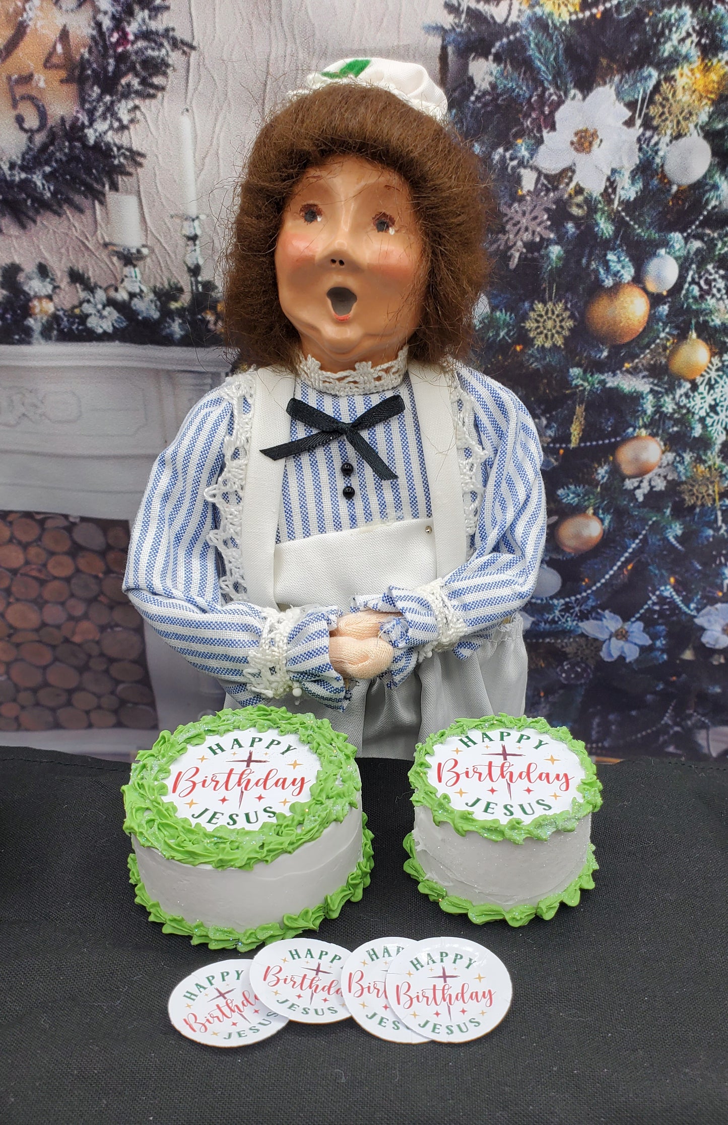 Figurine holding two birthday cakes with 'Happy Birthday Jesus' text, against a decorative background.