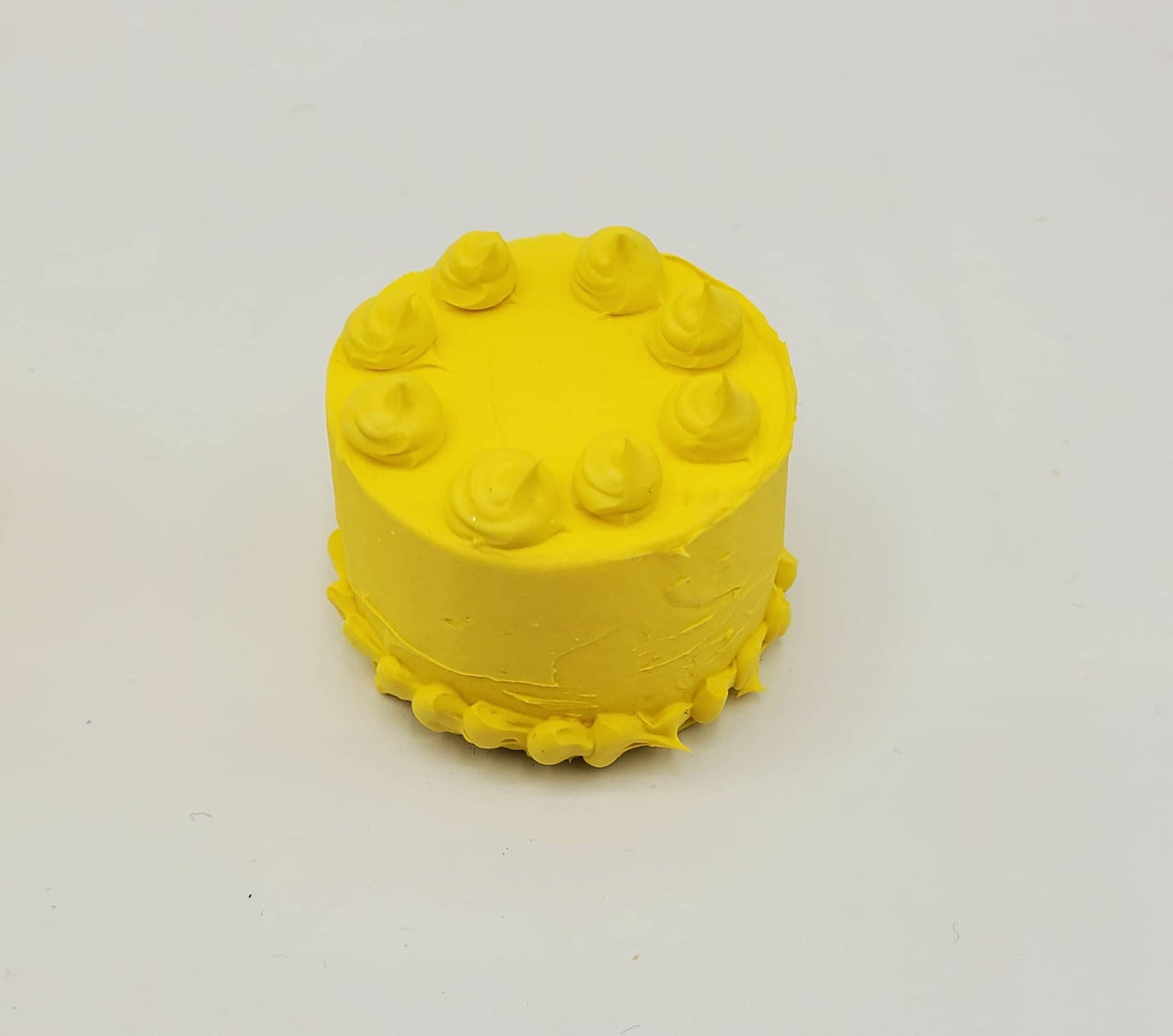 A small yellow cake-shaped object with piped frosting-like details on top and around the base, photographed on a plain light background.