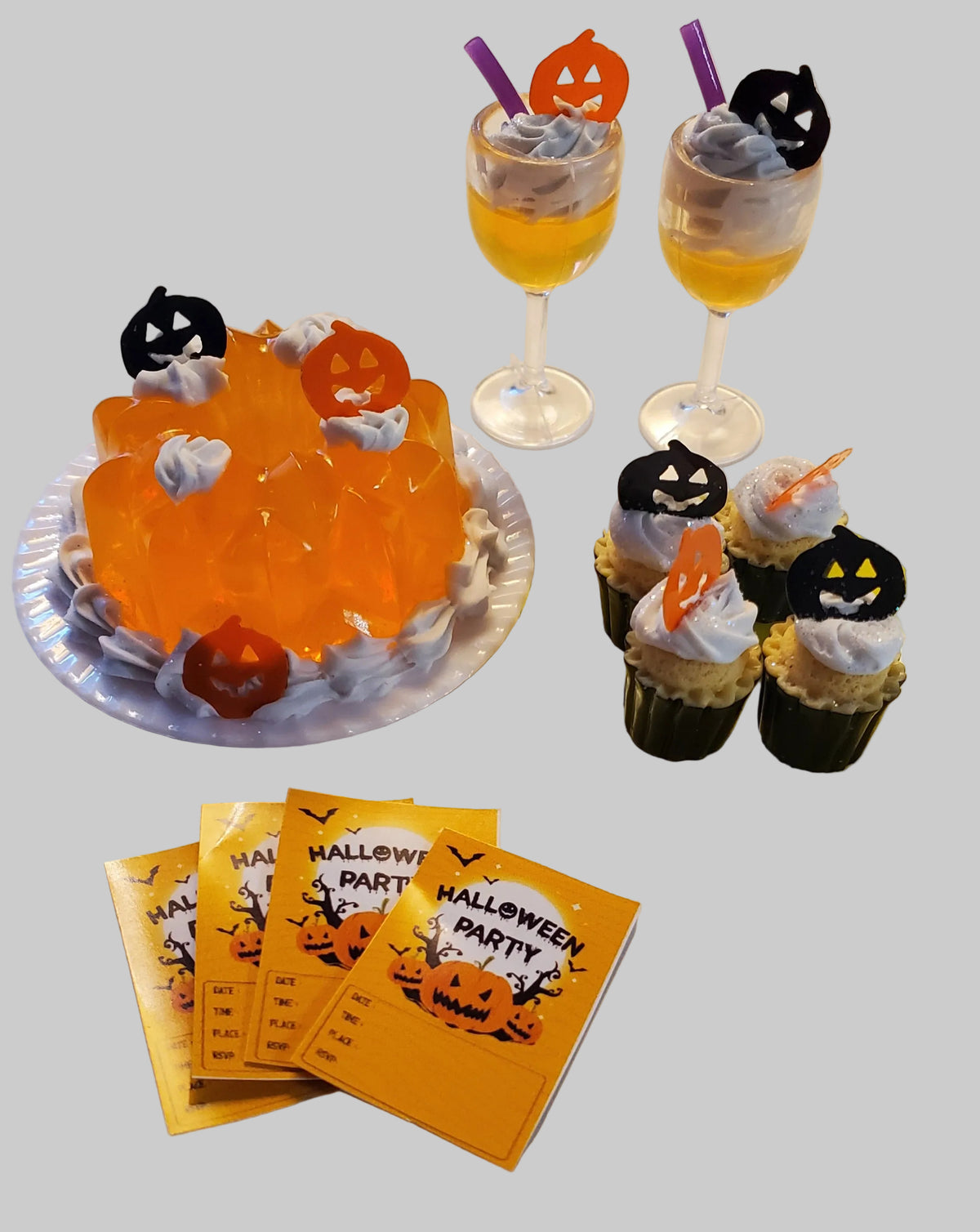 Halloween party food setup on a white table, including an orange jelly cake with jack-o'-lantern toppers, two glasses of yellow drink with whipped cream and purple straws, four cupcakes with white frosting and black pumpkin toppers, and several orange 'Halloween Party' tickets.