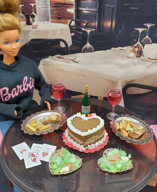 Barbie doll sitting at a table with heart-shaped cake, wine glasses, and food in a restaurant setting.