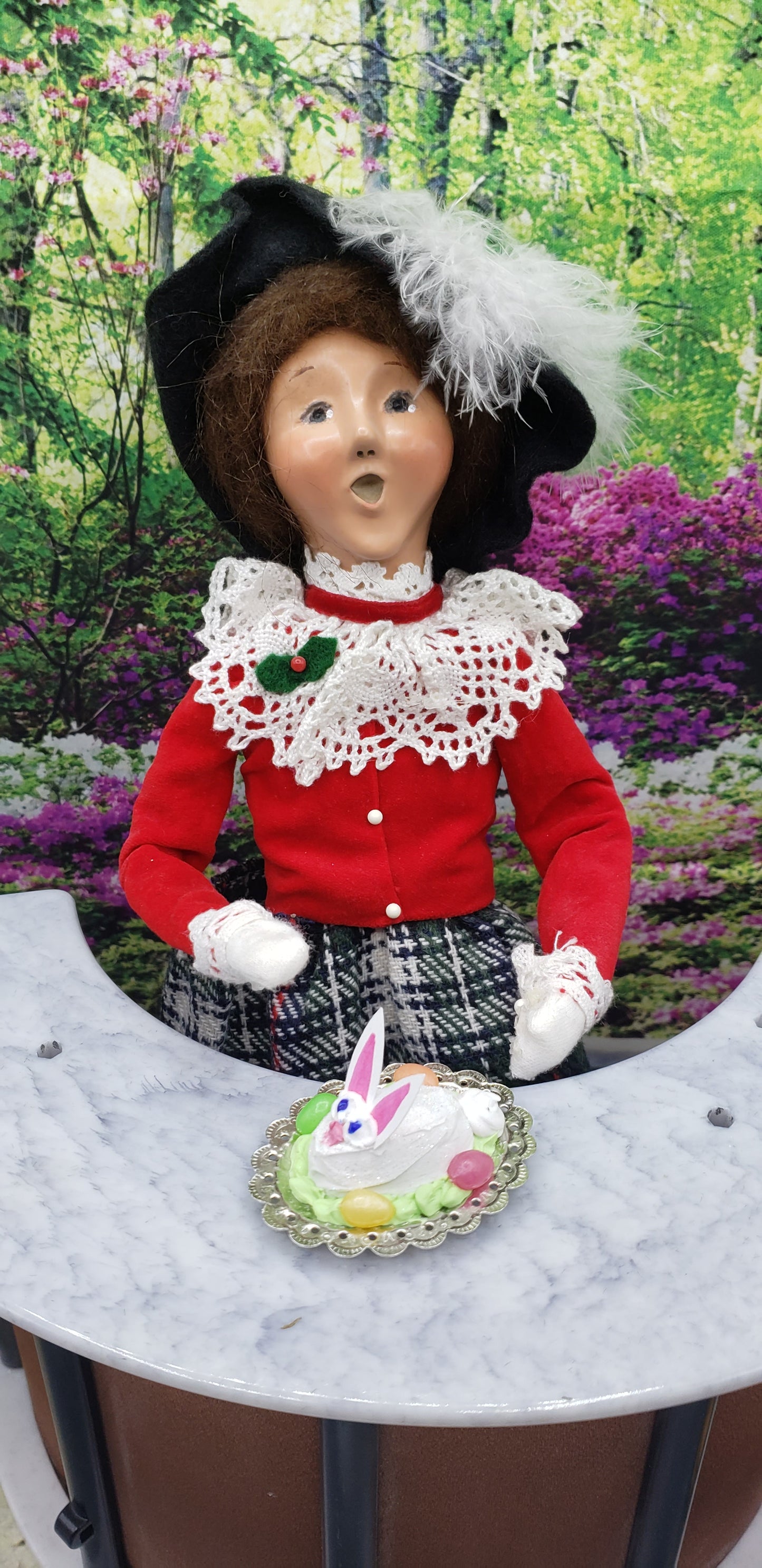 Fashion Doll size Easter bunny cake on a plate in front of a doll wearing red and black outfit with white lace collar