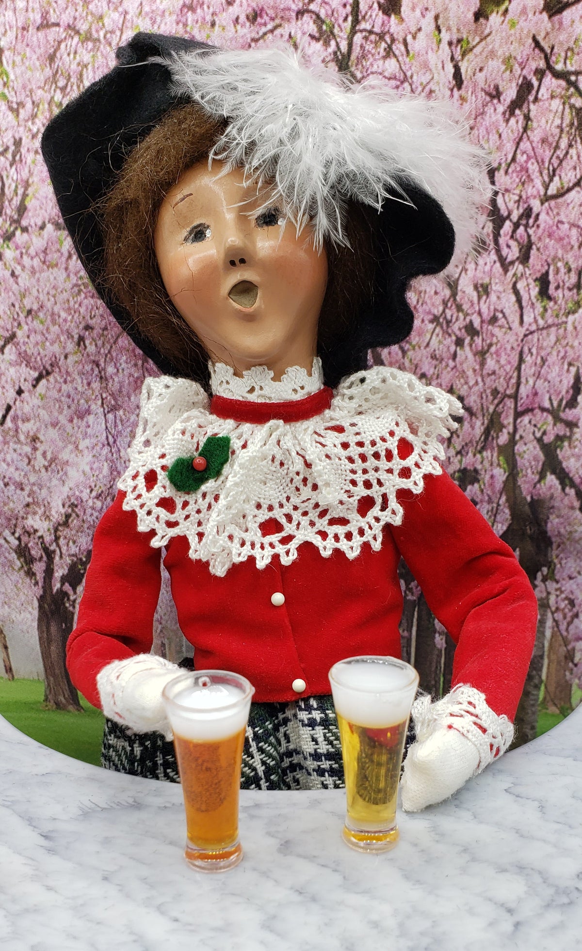 Two miniature beer mugs for dolls on a table beside a doll in a festive outfit