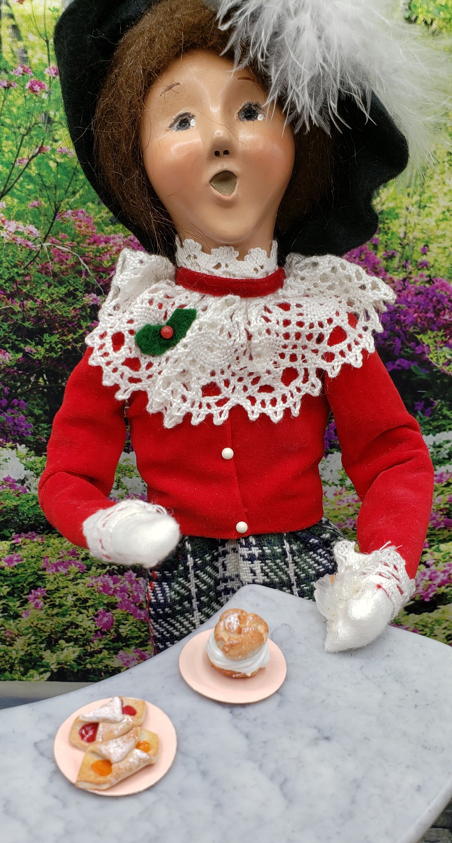 A miniature koloche doll food setup featuring a doll in a red outfit with pastries on small plates