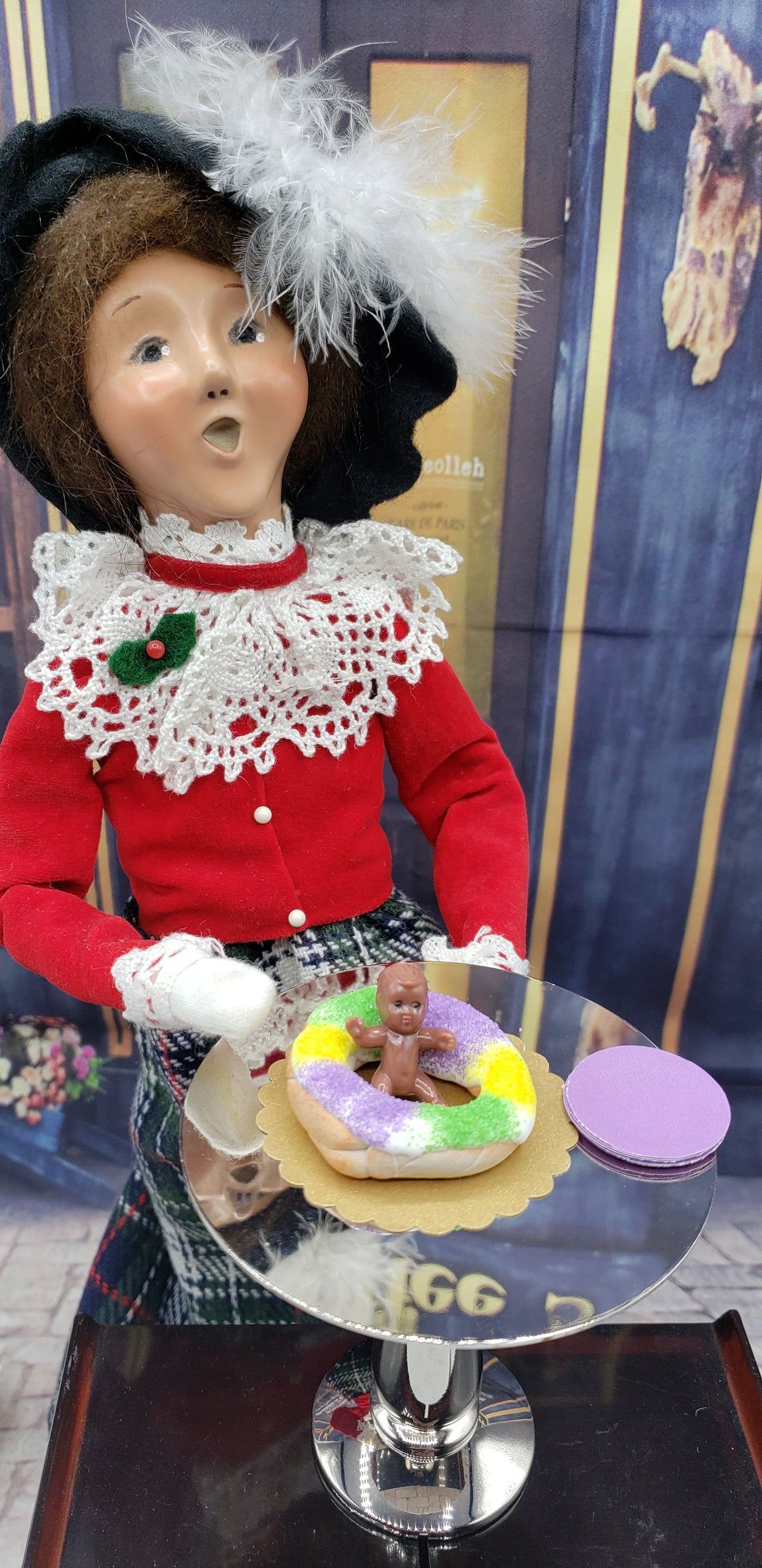 Doll in vintage attire with a miniature Mardi Gras king cake on a silver table