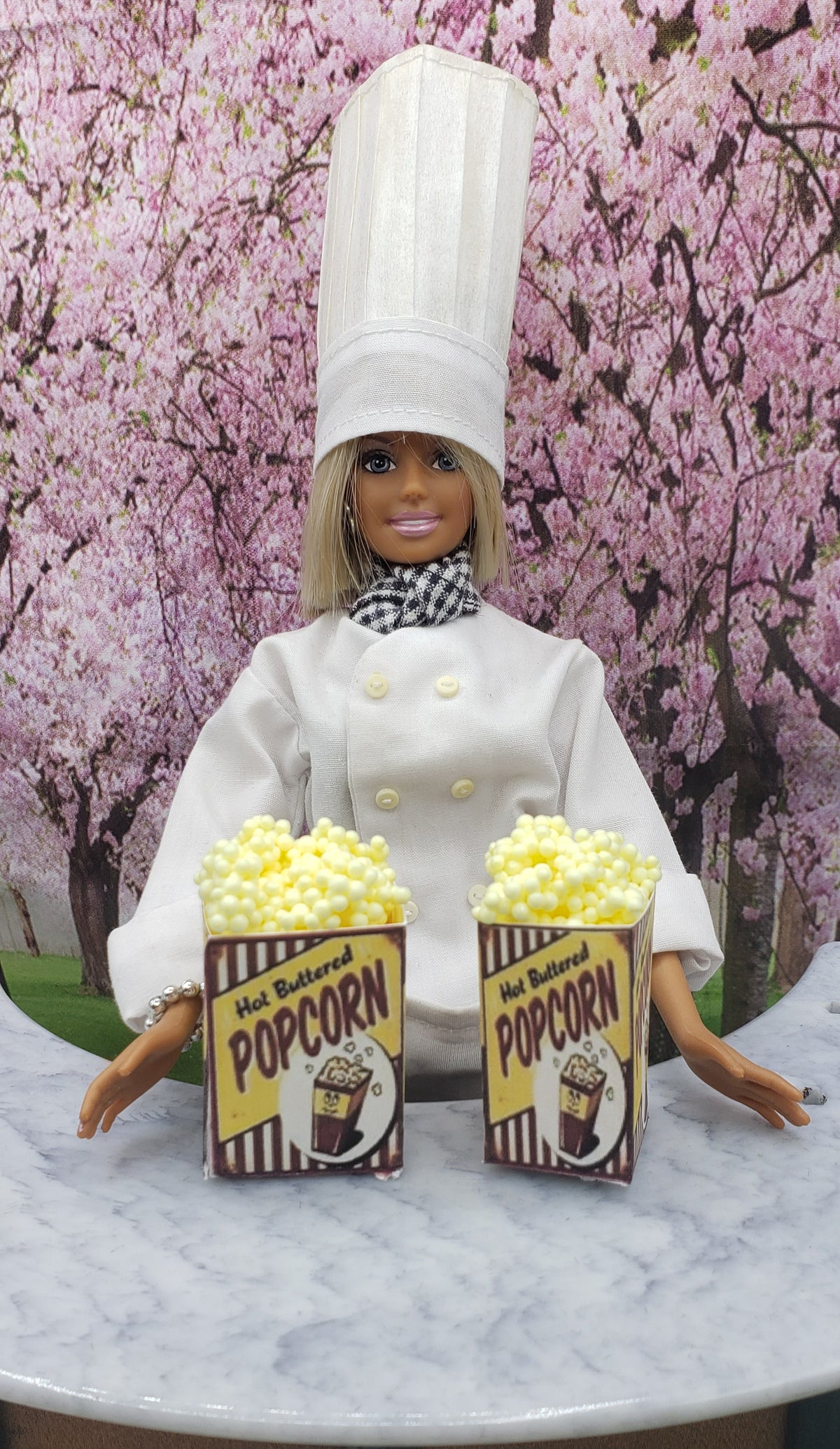 Chef doll in white uniform holding two miniature popcorn tubs with hot buttered popcorn label