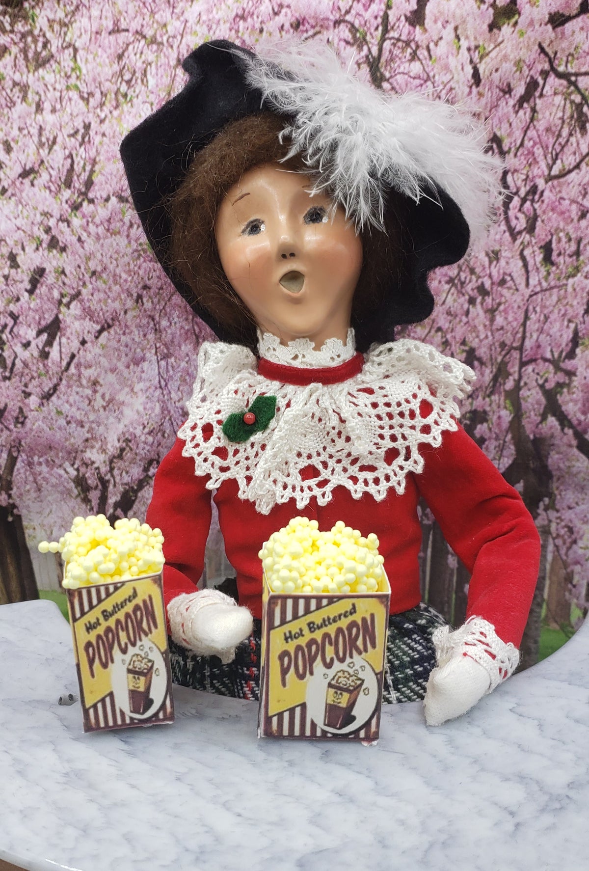 Chef Gina's miniature popcorn tub held by a carnival doll dressed in red with lace collar and feathered hat