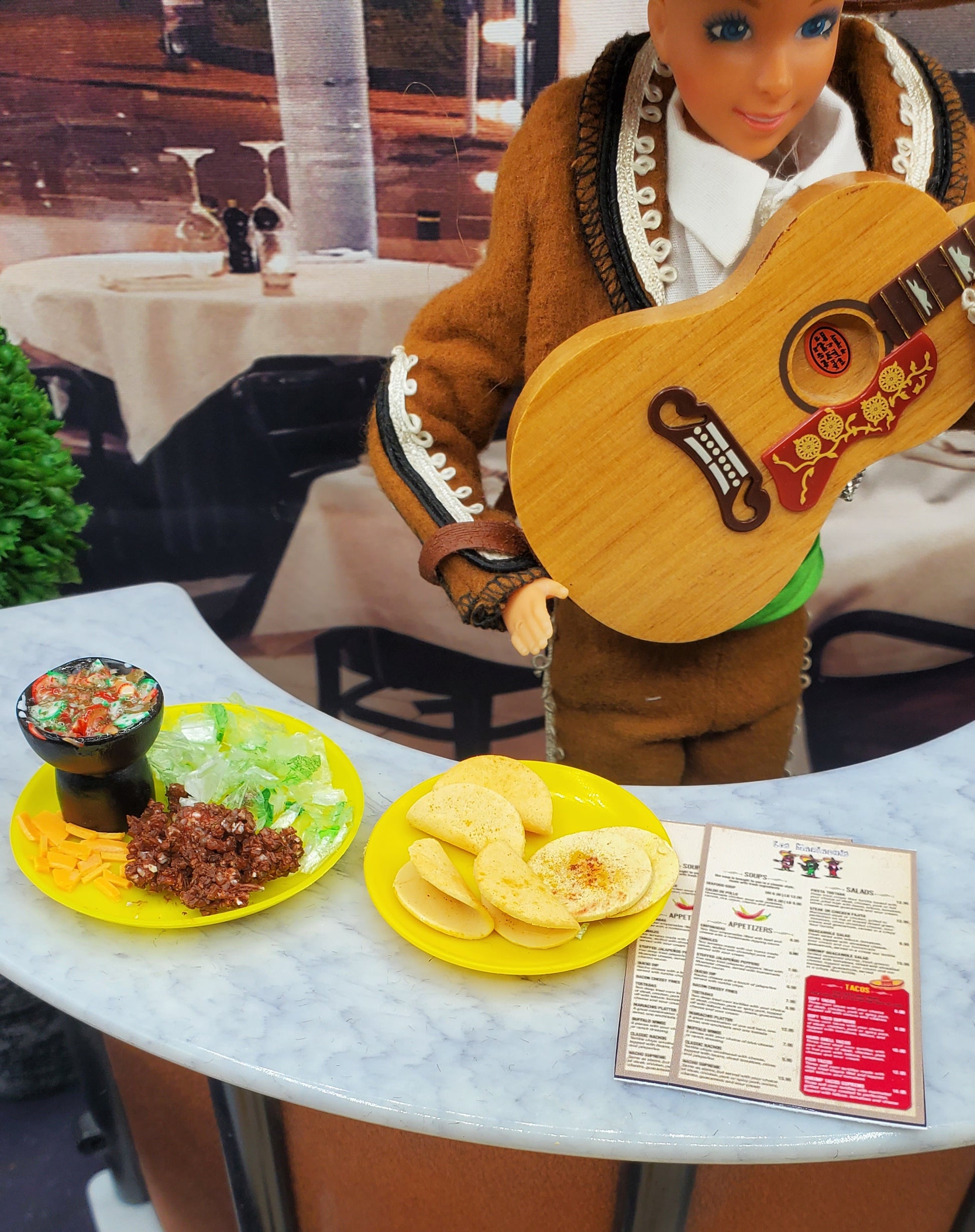 Barbie doll in traditional outfit with guitar, food on plates, and menu at a restaurant table.