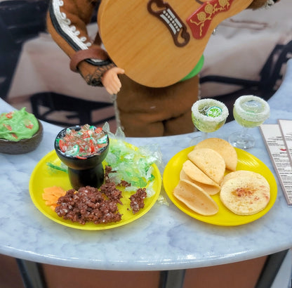Miniature plates with food items on a table, with a person in a costume in the background.
