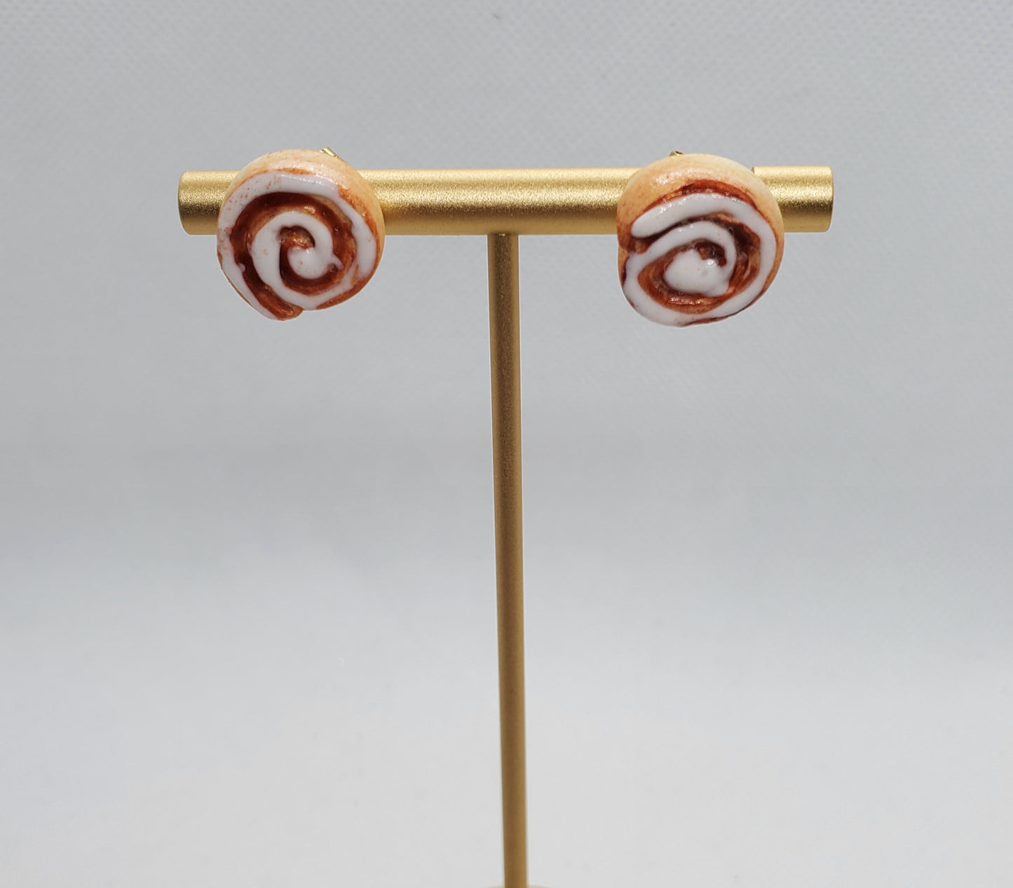 Chef Gina's miniature cinnamon roll earrings displayed on a gold T-stand with white icing detail