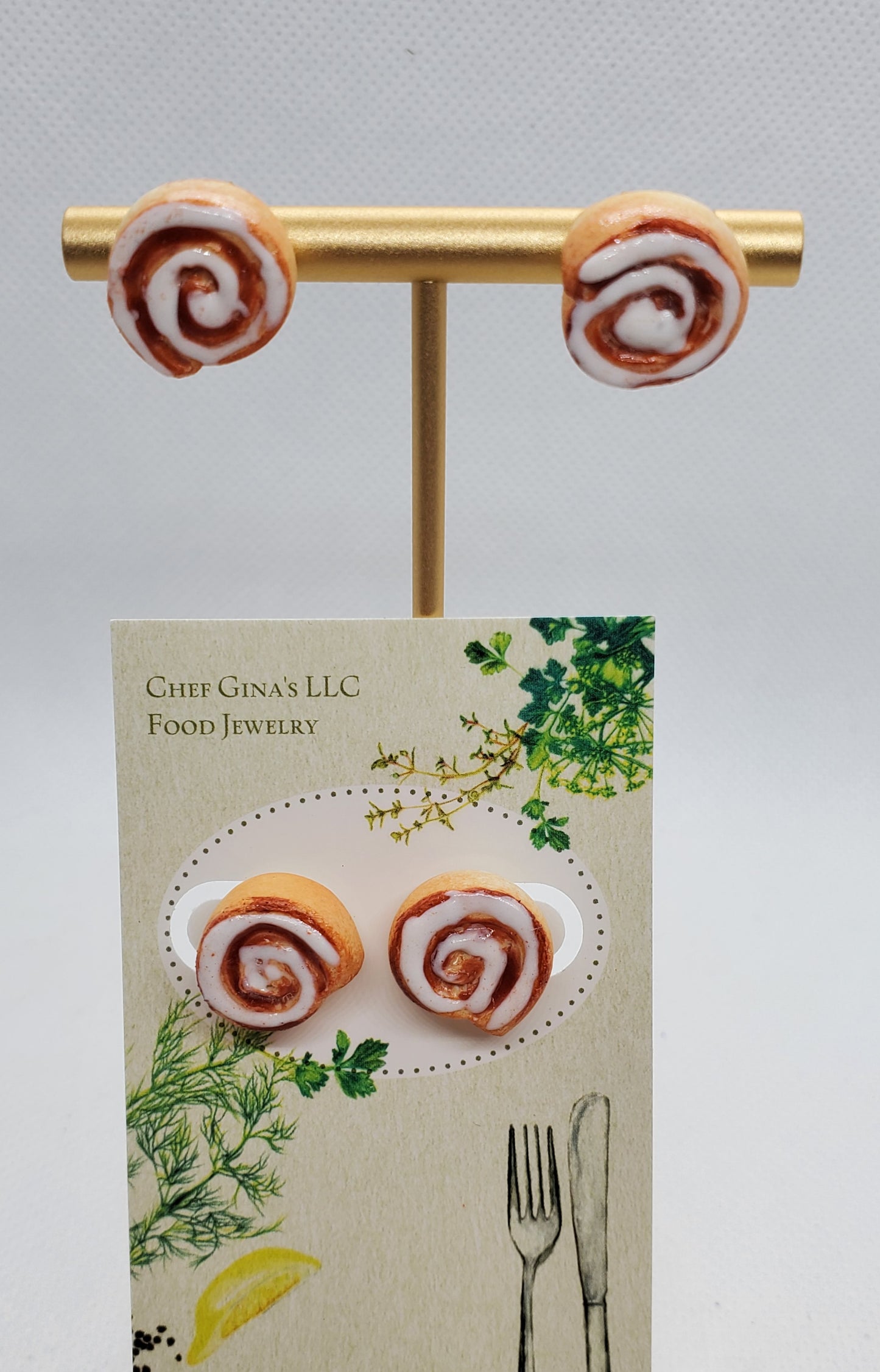 Chef Gina's miniature cinnamon roll earrings displayed on a gold stand with decorative packaging background
