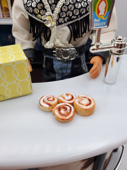 Miniature Texas cinnamon rolls for Fashion dolls displayed on a white counter beside a yellow box and doll figure