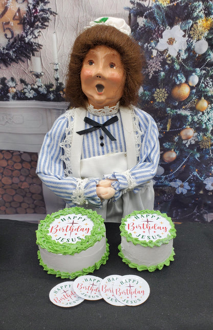 Figurine holding two birthday cakes with 'Happy Birthday Jesus' text, against a decorative background.