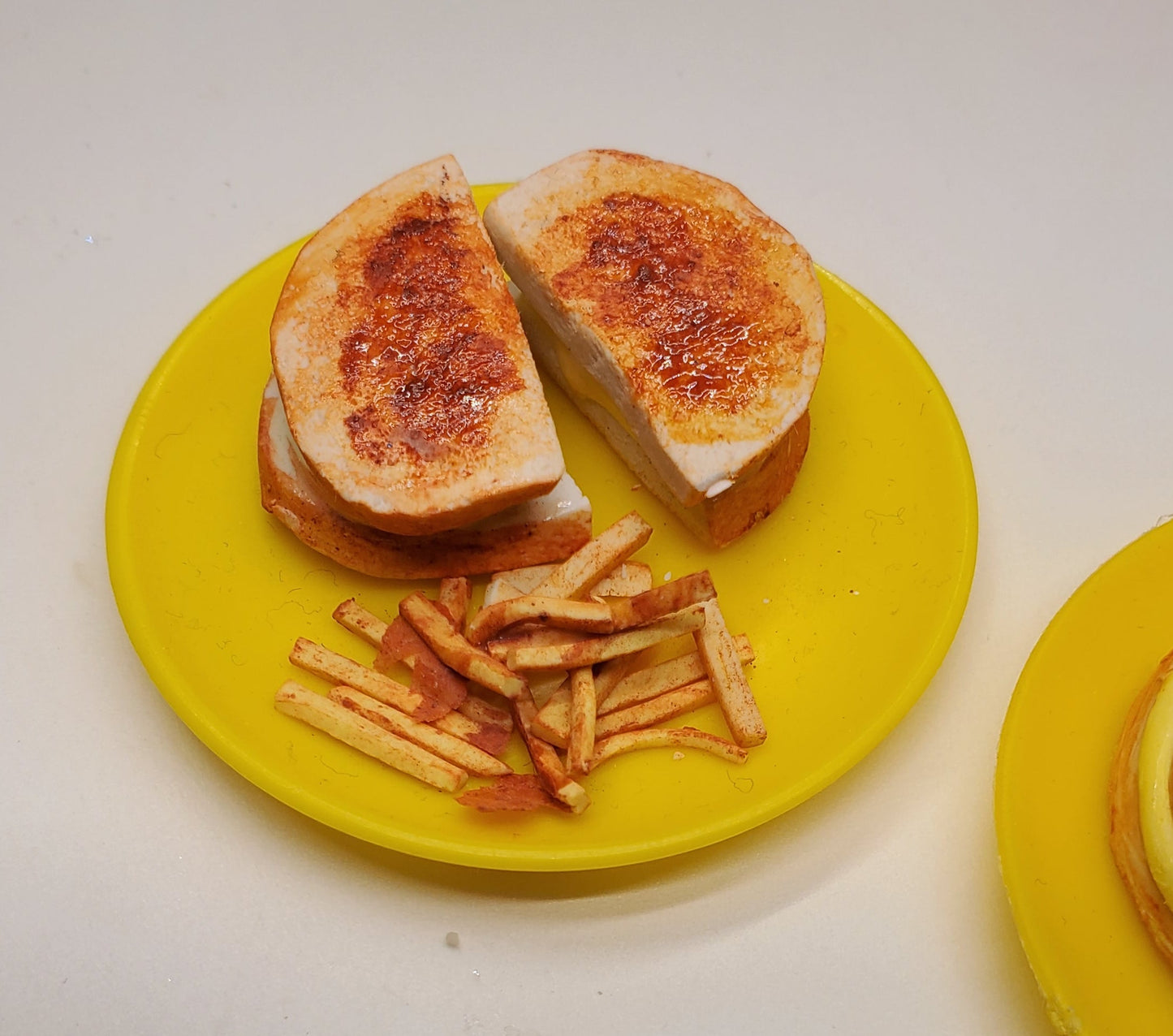 A miniature toy model of a toasted sandwich cut in half with french fries on a yellow plate, set on a light beige surface.