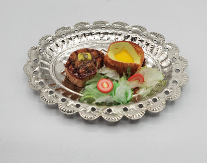 A decorative silver metal serving platter with a scalloped edge, holding a cooked food dish with a halved egg, two small browned pieces, and a small salad garnish on a light gray background.