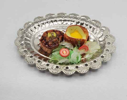 A decorative silver metal serving platter with a scalloped edge, holding a cooked food dish with a halved egg, two small browned pieces, and a small salad garnish on a light gray background.