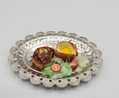 A scalloped silver metal plate holding two seared meat medallions, a halved baked potato with a bright yellow yolk, and a small salad photographed on a light gray background.