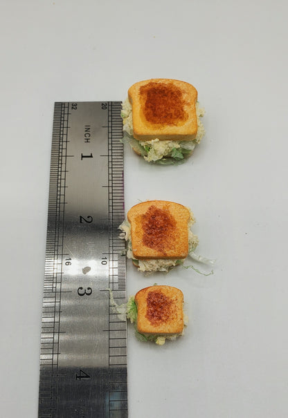 Three miniature sandwich slices with toasted bread and green filling arranged vertically next to a metal ruler on a white background.