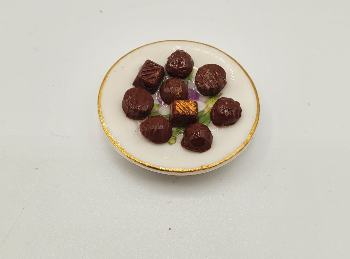 A miniature ceramic plate with gold trim holding several small dark brown chocolates, photographed on a plain white background.
