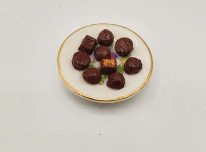 A miniature ceramic plate with gold trim holding several small dark brown chocolates, photographed on a plain white background.