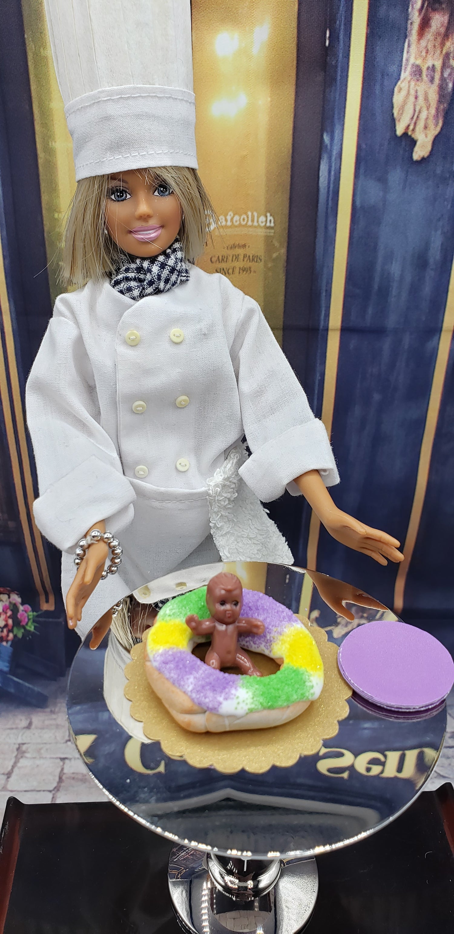 Miniature Mardi Gras king cake displayed on a cake stand with a chef doll in a white outfit
